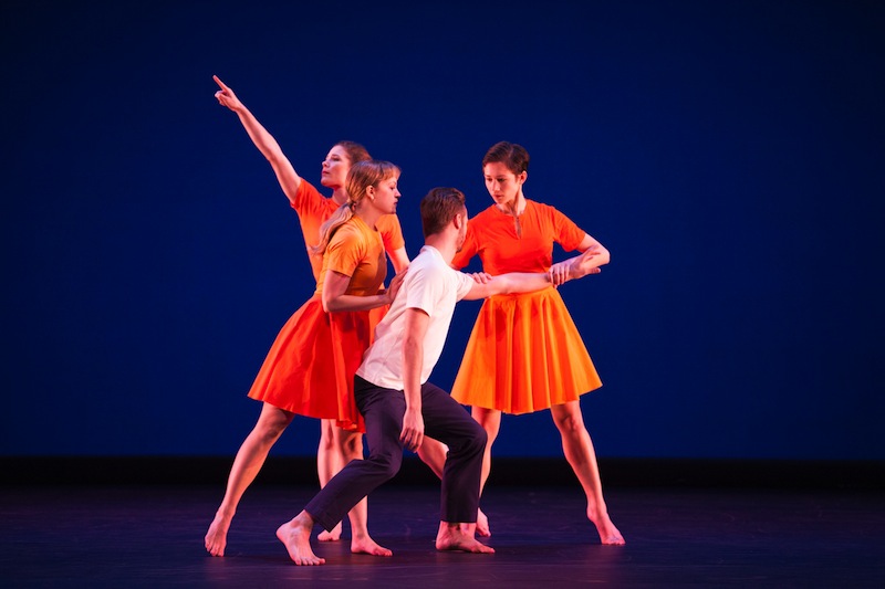 Three women in orange dresses cluster around a man, one pulls his arm while looking intently at him